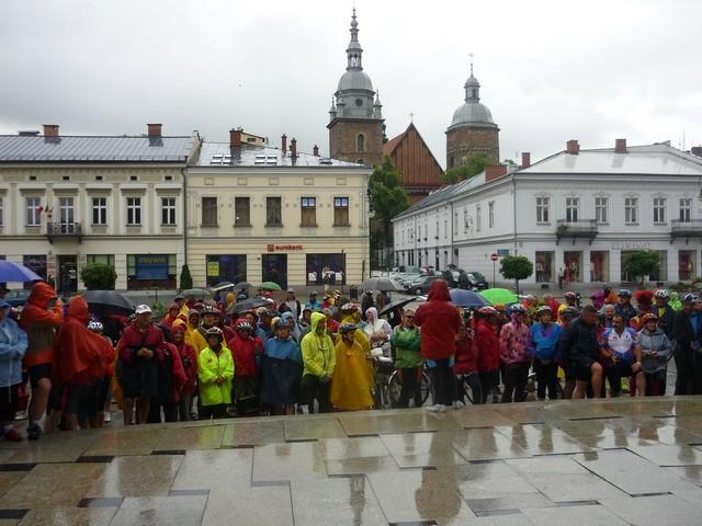 Na pierwszym planie uczestnicy zlotu w strugach deszczu, na drugim wieże bazyliki kolegiackiej
