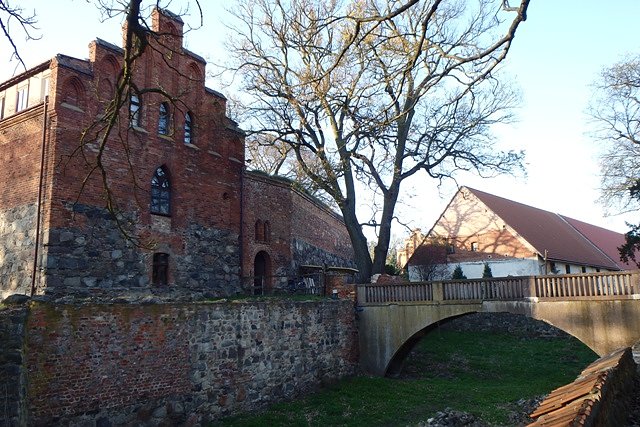 Na pierwszym planie - front skrzydła zachodniego, fragment muru i mostek przez fosę zamkową, na drugim - zabudowania gospodarcze zamku