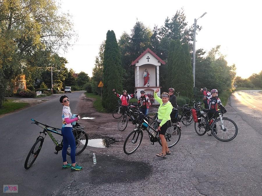 Grupa rowerzystów na wiejskiej drodze przy kapliczce